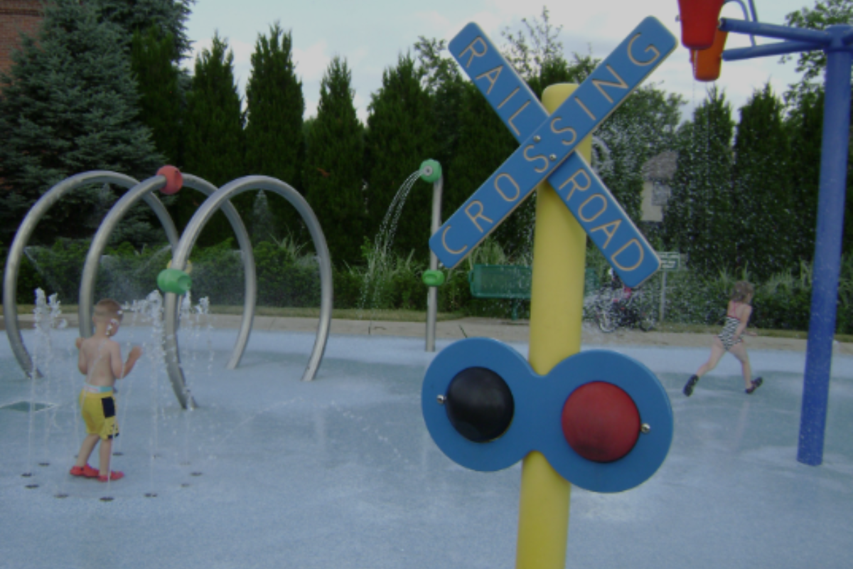 Railroad-themed splash pad at Village Green Park featuring a crossing sign and water arches in Delaware, Ohio.