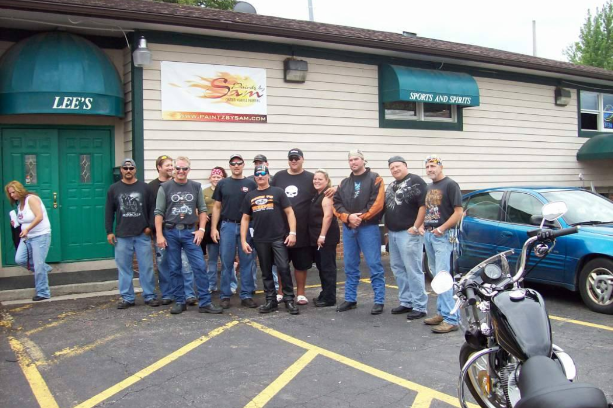 A group of people standing outside Lee's Sports and Spirits with motorcycles parked nearby.